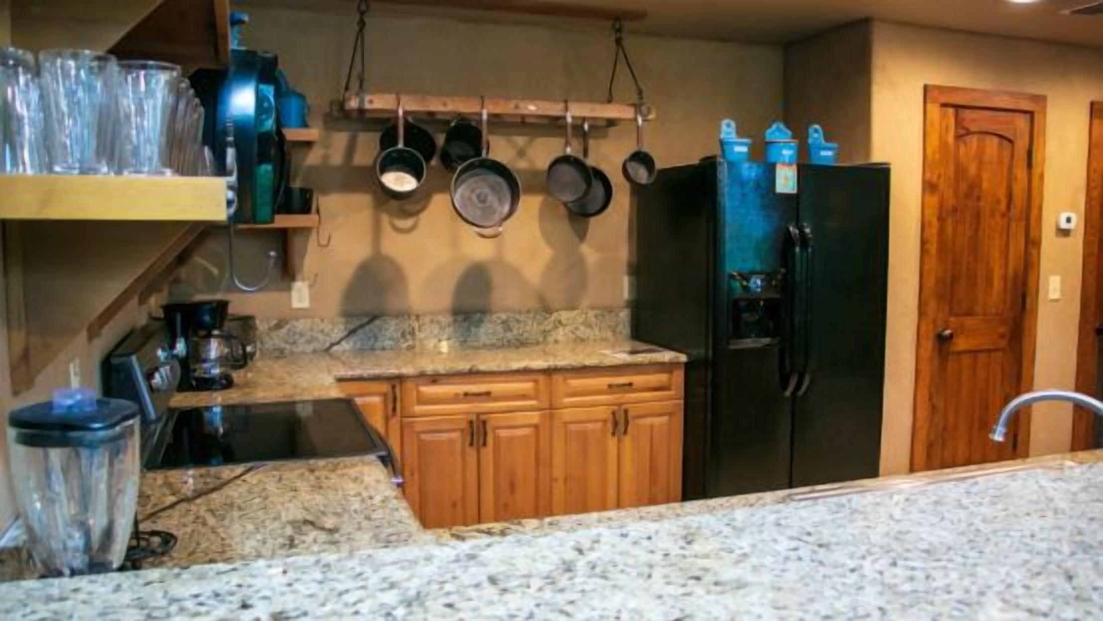 A kitchen with granite countertops, wooden cabinets, a black refrigerator, and hanging pots.