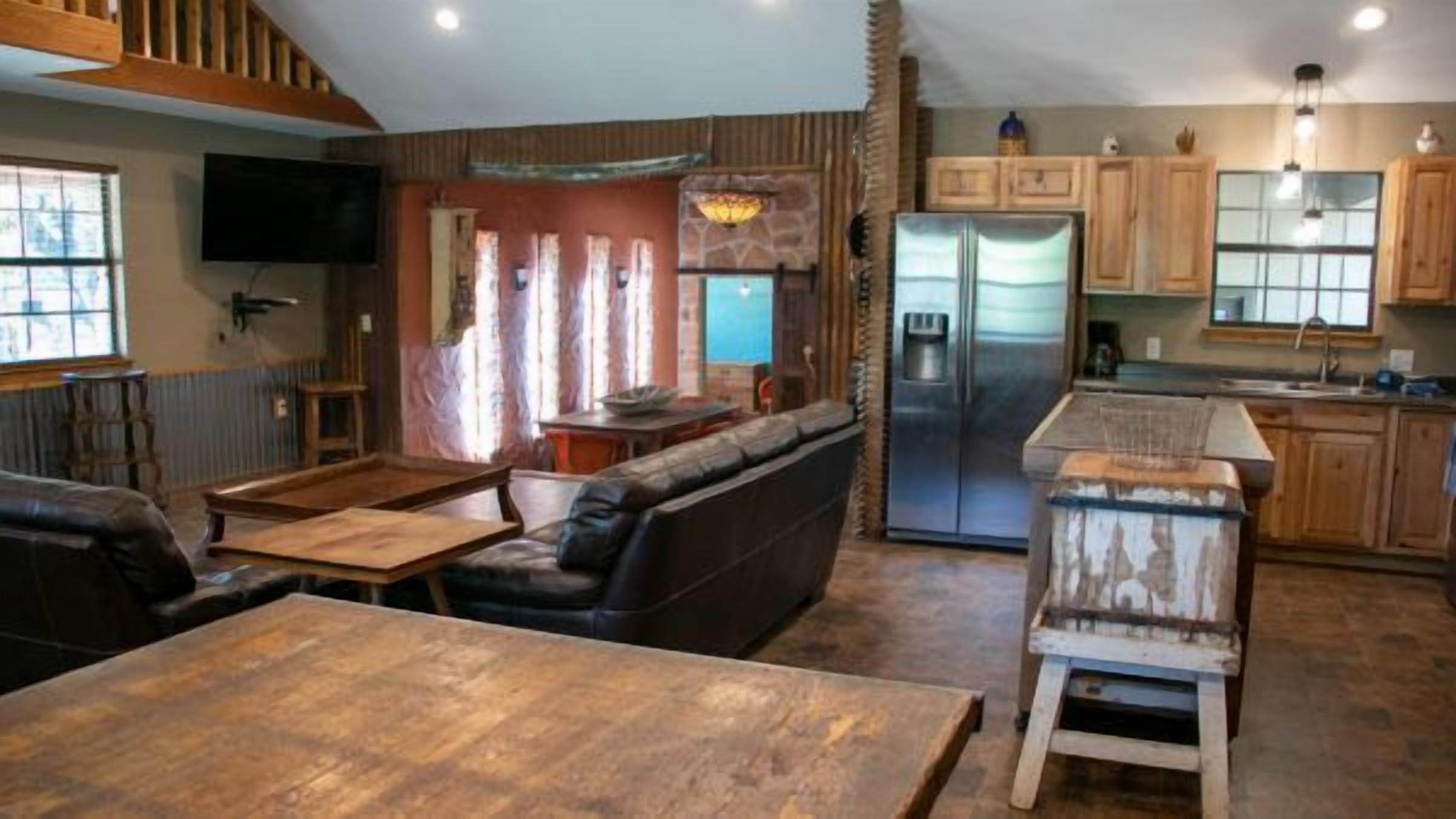 A rustic interior with a wooden dining table, leather sofa, and a kitchen area with wooden cabinets and a refrigerator.