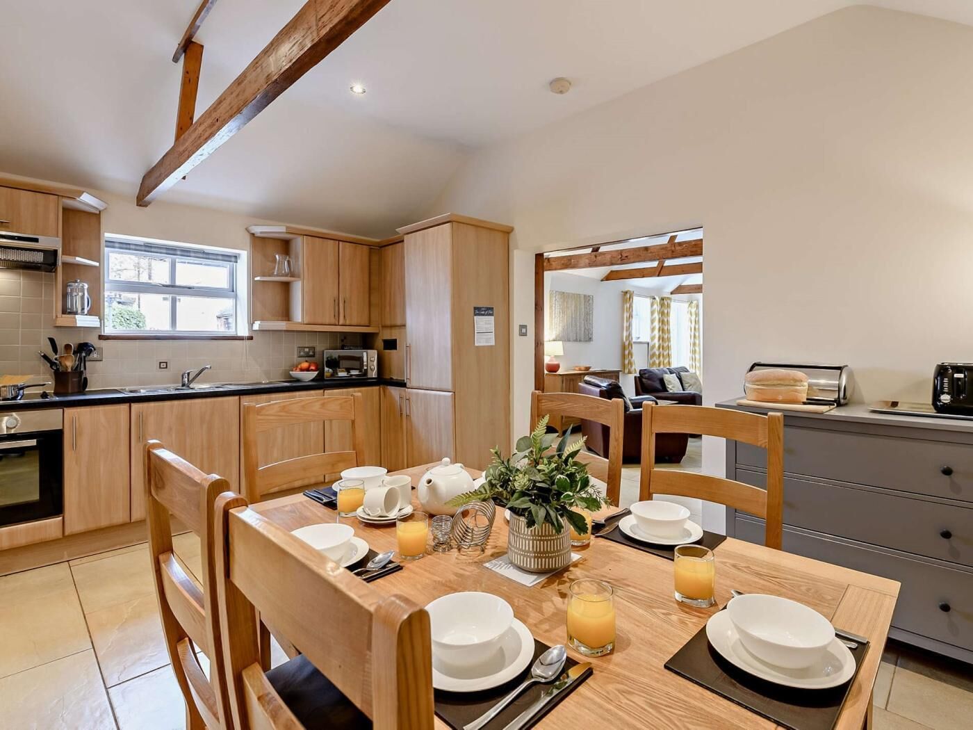 A modern kitchen with wooden cabinets, a dining table set for breakfast, and a view into the living area.