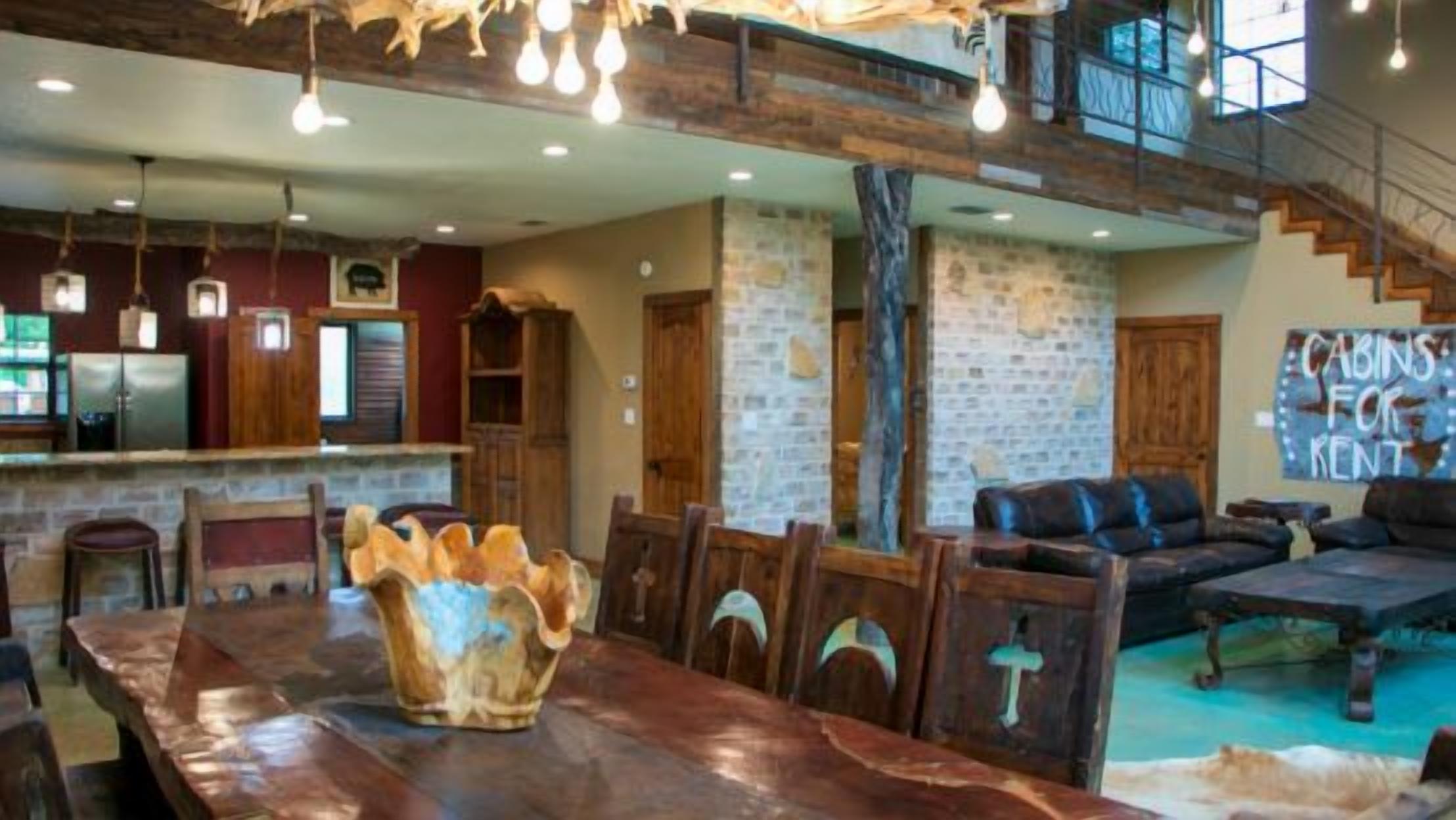 A spacious dining area with a long wooden table and chairs, a rustic stone wall, and a staircase leading to an upper level.