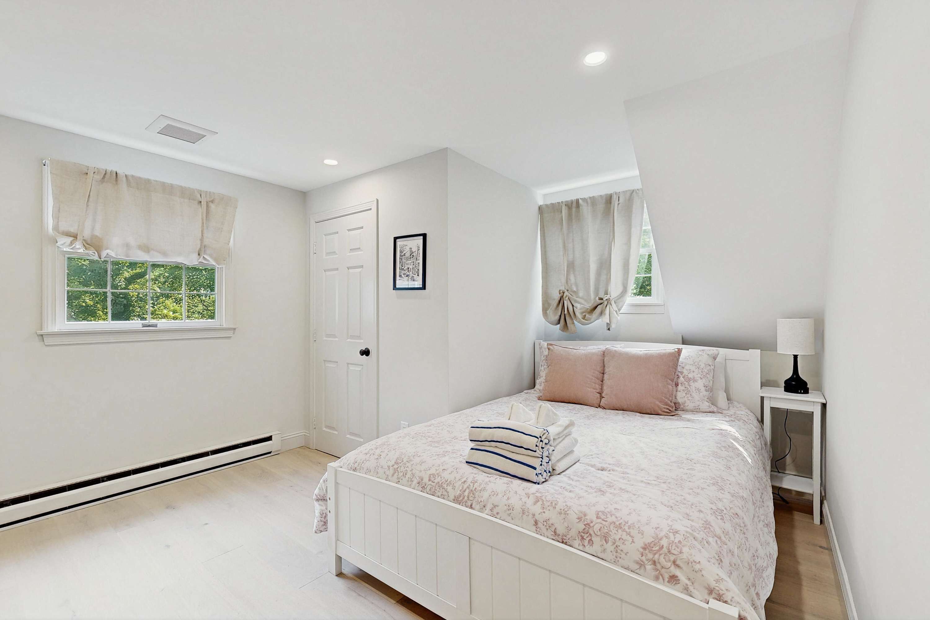 A bedroom with a bed, two windows with curtains, a door, a bedside table with a lamp, and a picture on the wall.