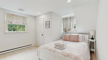A bedroom with a bed, two windows with curtains, a door, a bedside table with a lamp, and a picture on the wall.