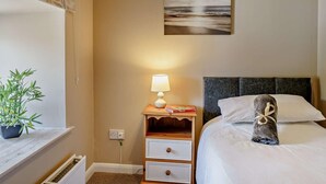 A hotel room with a bed, bedside lamp, a nightstand with a book, a plant, and a painting on the wall. - 3 Bed in Cosheston (oc-90732) (Cosheston)