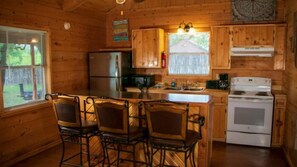 A wooden cabin interior with a kitchen area, a dining table with chairs, and a large window. - Riverfront Cabins  and  Vacation Homes (Rio Frio)