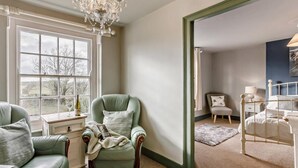 A room with a large window, a green armchair, a white bedside table, and a bed with a white headboard.