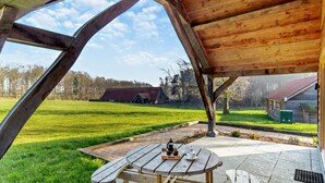 Een overdekt buitengebied met een picknicktafel en banken, een uitzicht op een grasveld en een gebouw in de verte.