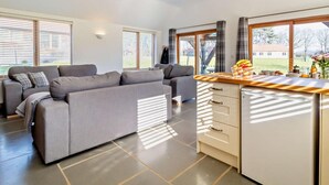 Een moderne woonkamer met een grijze bank, een keuken met een houten aanrecht en grote ramen met zonwering.