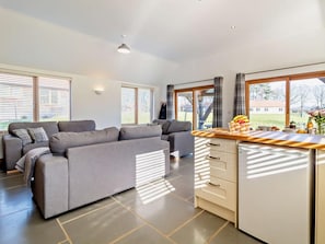 Ein modernes Wohnzimmer mit grauem Sofa, einer Küchenzeile mit Holzarbeitsplatte und großen Fenstern mit Jalousien.