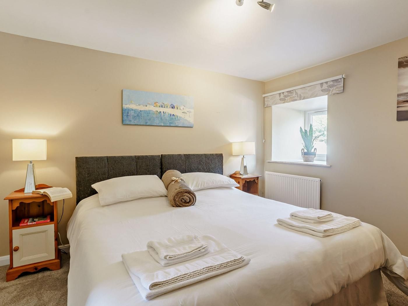 A hotel room with a bed, two bedside tables, a window with a plant, and a painting on the wall.