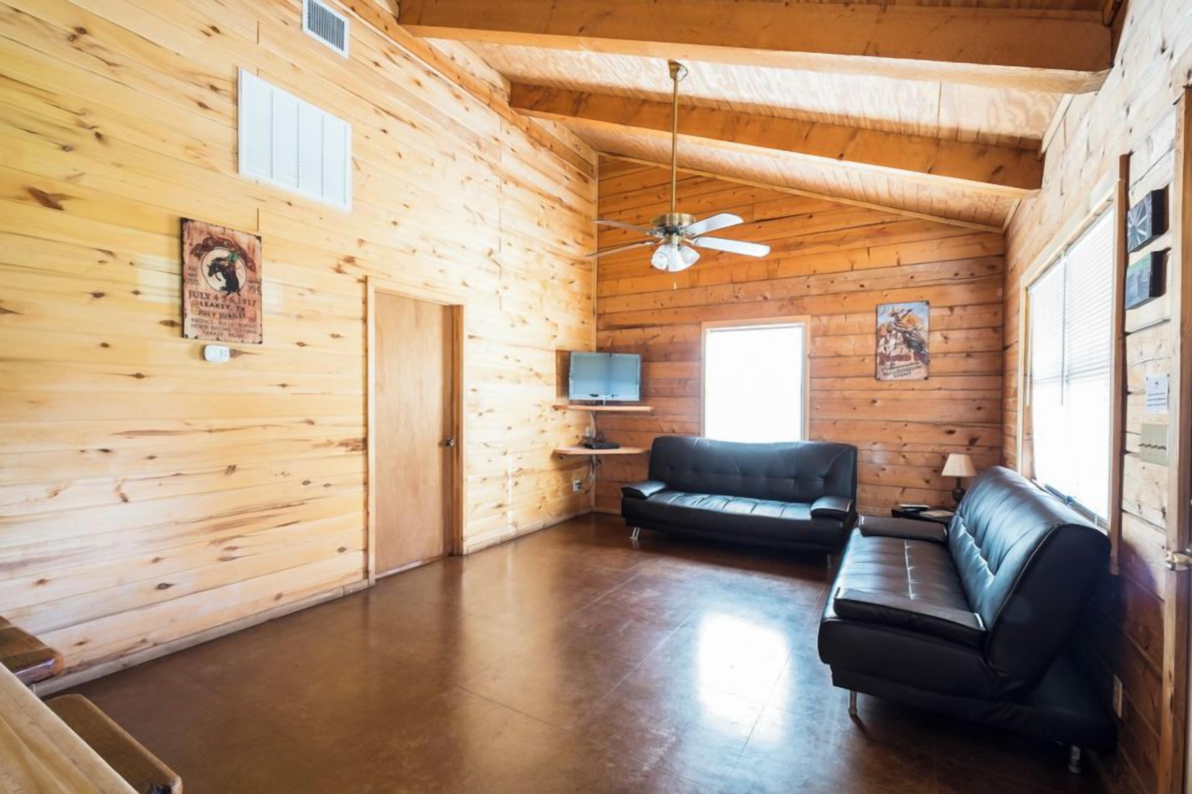 Ein Blockhaus-Innenraum mit Deckenventilator, einem schwarzen Ledersofa, einem Fernseher auf einem Standfuß und Holzwänden.