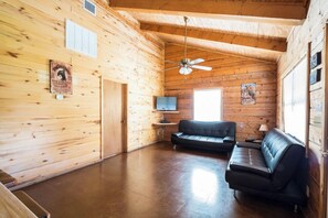 Ein Blockhaus-Innenraum mit Deckenventilator, einem schwarzen Ledersofa, einem Fernseher auf einem Standfuß und Holzwänden.