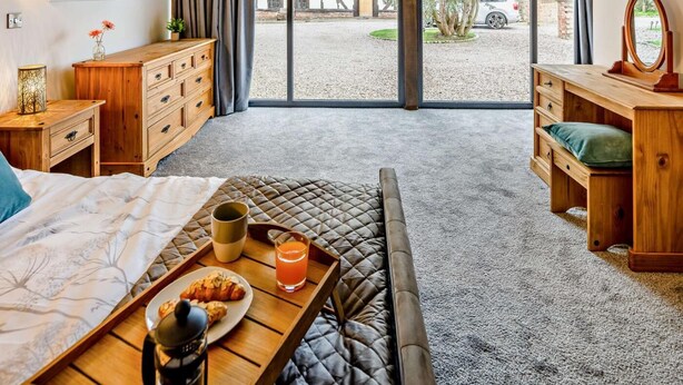 A bedroom with a bed, a wooden dresser, a bedside table with a lamp, a window with curtains, and a view of the outside. - 1 Bed in Northwich (oc-91435) (Northwich)
