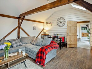 A cozy loft-style living space with exposed wooden beams, a grey sofa, a red plaid blanket, and a dining area with a wooden table and chairs. - 1 Bed in Northwich (oc-91435) (Northwich)