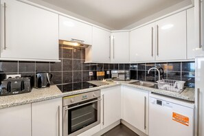 A modern kitchen with white cabinets, a black tiled backsplash, a built-in oven, and a dishwasher. - 2 Bed in Brixham (oc-91427) (Brixham)