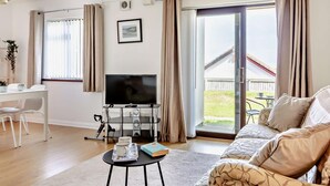 A modern living room with a sofa, a coffee table with a book and mug, a TV, and a dining area with chairs and a table. - 2 Bed in Brixham (oc-91427) (Brixham)
