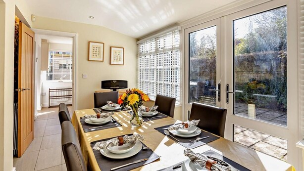 Un coin repas avec une table dressée pour quatre personnes, un vase rempli de fleurs et une vue sur le jardin grâce à de grandes fenêtres.