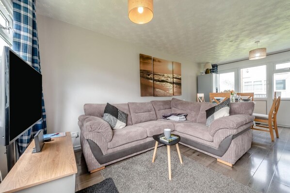 A modern living room with a large sectional sofa, a flat-screen TV, a wooden coffee table, and a dining area with chairs and a table. - 2 Bed in Freshwater East (oc-91445) (Freshwater East)