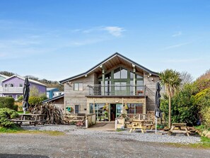 A two-story wooden cabin with a balcony, a gravel driveway, and picnic tables. - 2 Bed in Freshwater East (oc-91445) (Freshwater East)