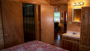 Una habitación de madera con una cama, un baño con lavamanos y espejo, y una puerta que da a otra habitación.