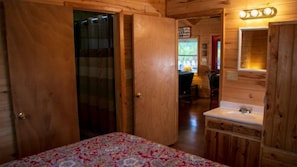 Ein Zimmer mit Holzboden, einem Bett, einem Badezimmer mit Waschbecken und Spiegel sowie einer Tür zu einem weiteren Raum.