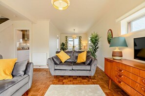 A modern living room with a grey sofa, wooden flooring, a white radiator, a wooden cabinet, and a television on the wall. - 3 Bed in Swanage (oc-91451) (Swanage)