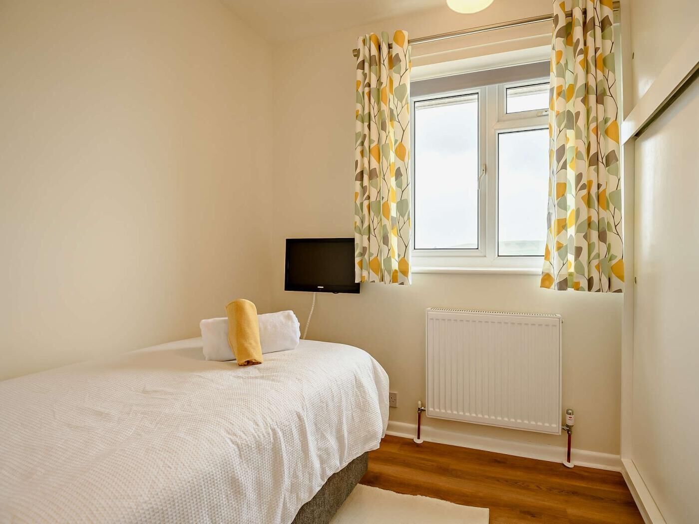 A bedroom with a bed, a television, a window with curtains, and a radiator.