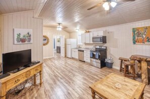 A room with a wooden floor, a television on a wooden stand, a dining table, and a kitchen area with cabinets and a microwave. - Riverfront Cabins  and  Vacation Homes (Rio Frio)