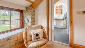 A corner room with a stone wall, a wicker chair with a cushion, a window, and a view of a kitchen. - 3 Bed in Tenby (oc-91524) (Sageston)