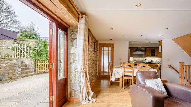 A cozy living room with a brown leather sofa, a dining table with chairs, and a kitchen in the background. - 3 Bed in Tenby (oc-91524) (Sageston)