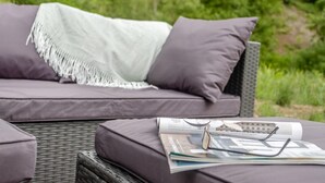 Un canapé en osier avec des coussins et une couverture, une paire de lunettes et un magazine sur une table.