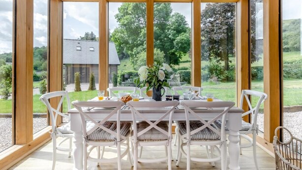 Une table de petit-déjeuner, avec des chaises, un vase de fleurs et une vue sur un paysage verdoyant.