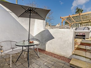 A patio with a round table, chairs, a grill, and a white wall. - 1 Bed in Barnstaple (oc-91612) (Bishop's Tawton)