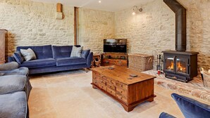 A stone-walled room with a blue sofa, wooden coffee table, and a wood-burning stove. - 4 Bed in Shaftesbury (oc-91456) (East Melbury)