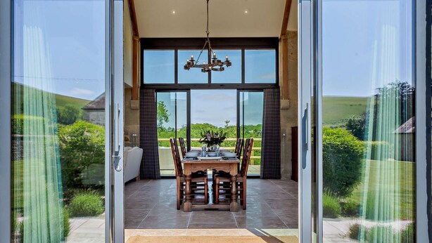 A modern house with a dining area, large glass doors, and a view of the countryside. - 4 Bed in Shaftesbury (oc-91456) (East Melbury)