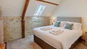 A bedroom with a stone wall, wooden beams, a skylight, a bed with white linens, and a nightstand with a lamp. - 4 Bed in Shaftesbury (oc-91456) (East Melbury)