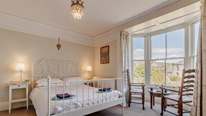 Ein Schlafzimmer mit einem Bett, zwei Nachttischen, einer Deckenleuchte, einem Fenster mit Vorhängen und Blick auf Gebäude.