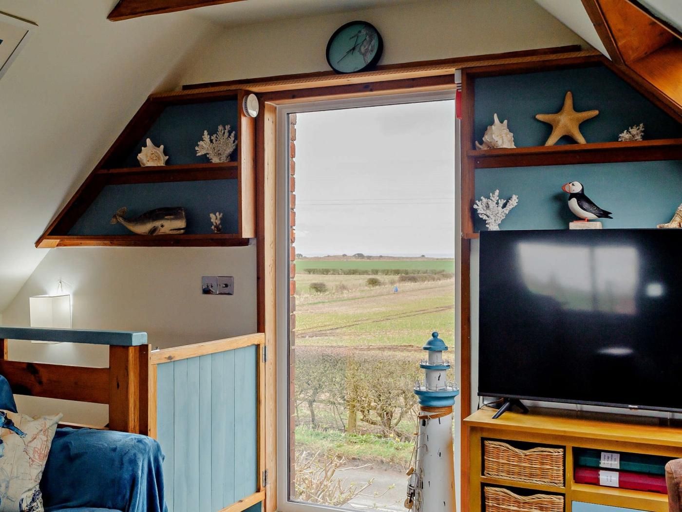 A cozy room with a television, wooden shelves with decorative items, a clock, and a view of the countryside.