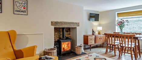 Una sala de estar acogedora con sofá, chimenea, zona de comedor con sillas, televisor montado en la pared y ventana con vistas.