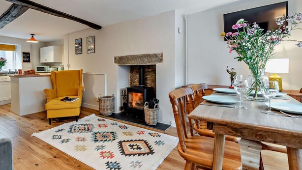 A cozy living room with a fireplace, a dining area with wooden chairs, a yellow armchair, and a kitchen in the background.