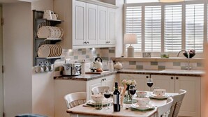 Una cocina con armarios blancos, una mesa de comedor lista para dos personas y un ventanal con persianas.