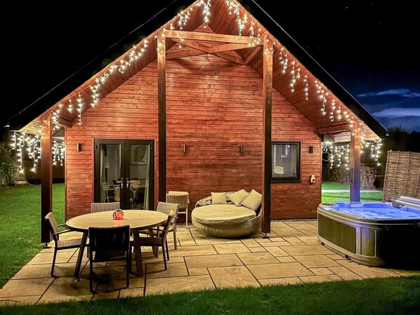 A wooden cabin with a hot tub, outdoor seating, and string lights.