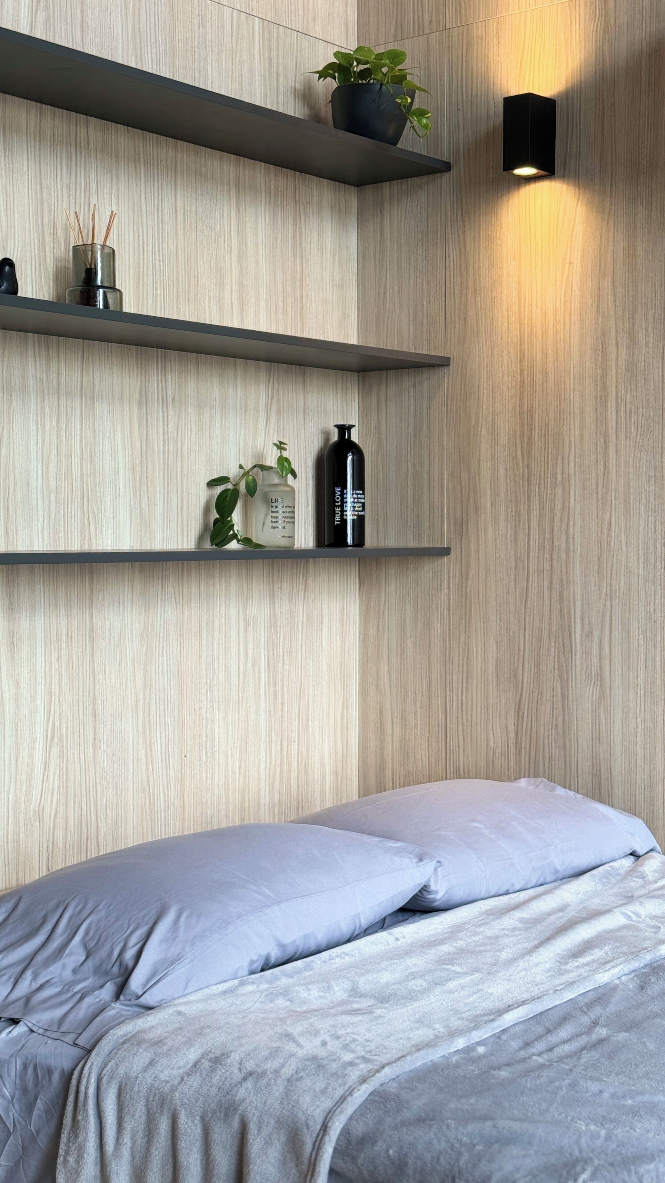A bedroom with a bed, a nightstand, and a wall-mounted shelf with a plant and a bottle.