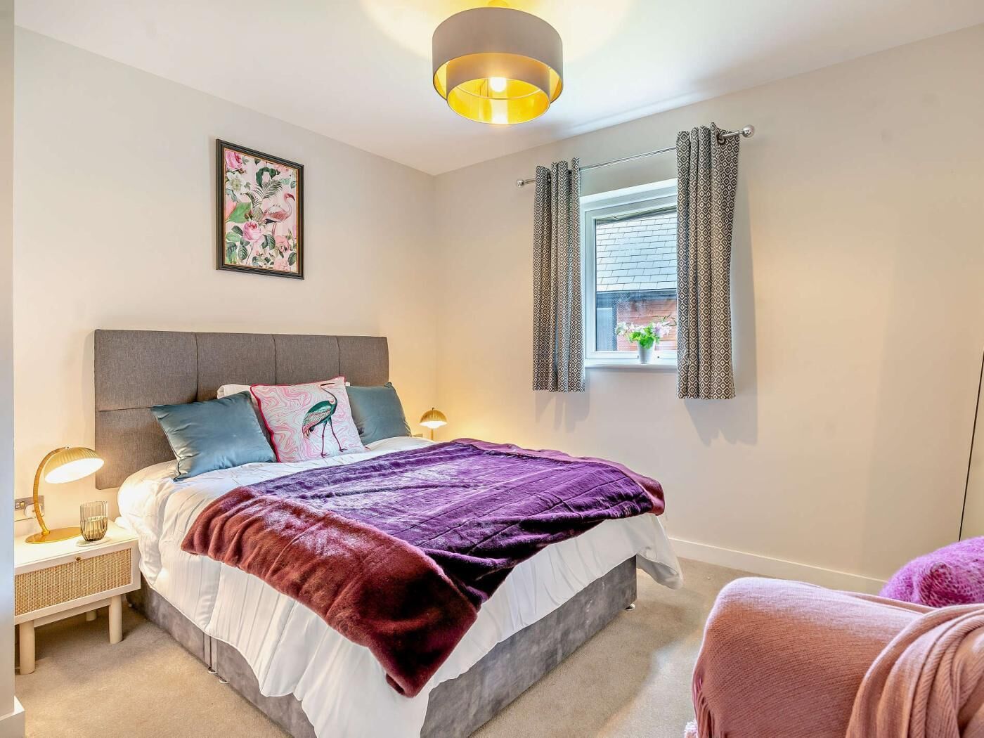 Une chambre à coucher comprenant un lit, des tables de chevet, une fenêtre avec des rideaux, un luminaire fixé au mur et un tableau encadré.