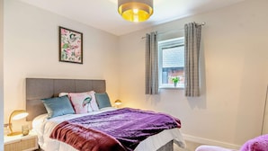 Ein Schlafzimmer mit einem Bett, Nachttischen, einem Fenster mit Vorhängen, einer Wandleuchte und einem gerahmten Bild.