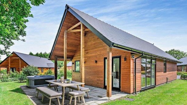 Eine moderne Holzblockhütte mit überdachter Terrasse, Whirlpool und gepflegtem Rasen.