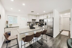 Et moderne køkken med hvide skabe, en central ø med barstole, rustfrie stålemmer og et bølgemønstret backsplash.