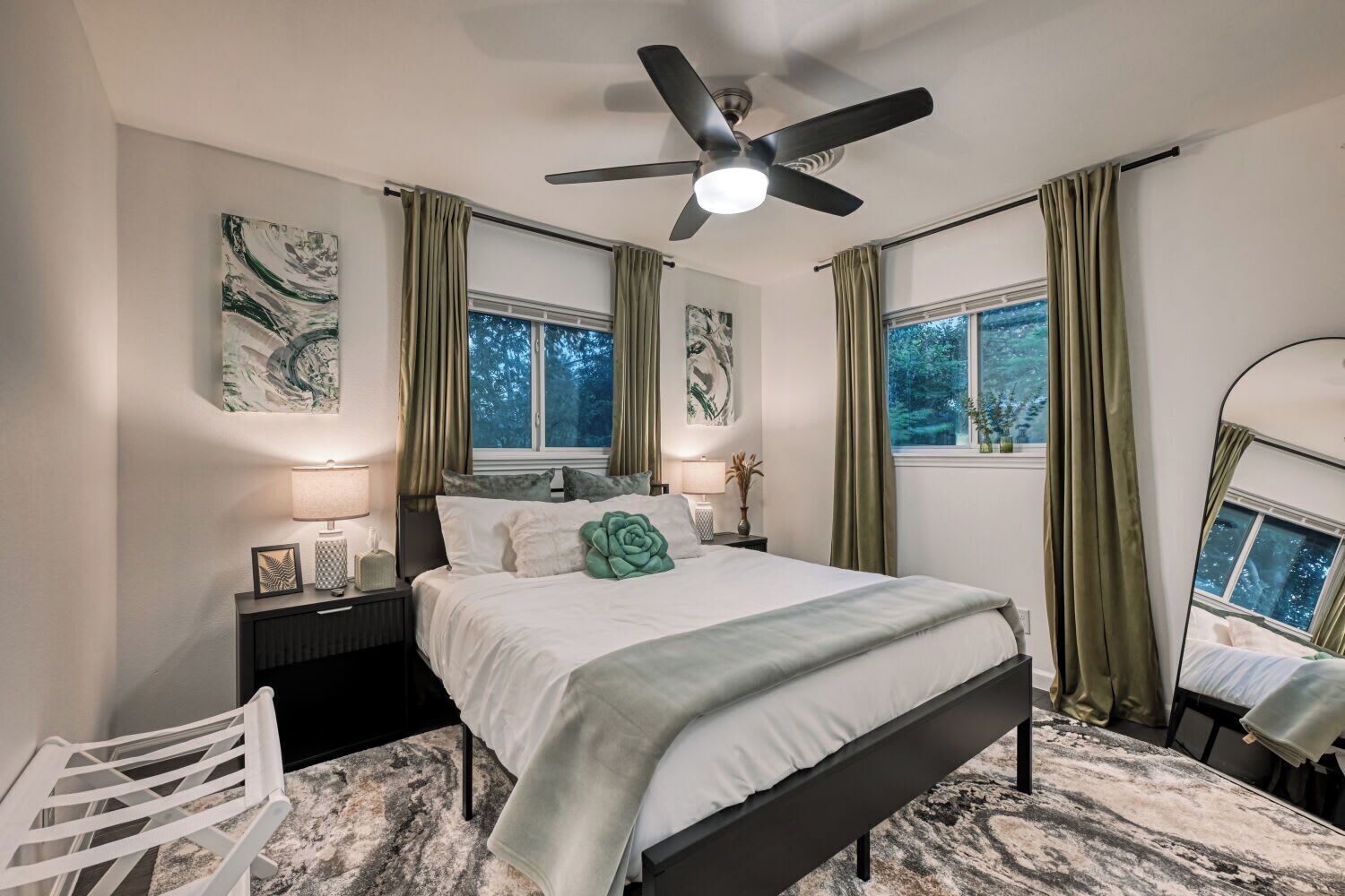 A bedroom with a bed, nightstands, a chair, a ceiling fan, and two windows with curtains.