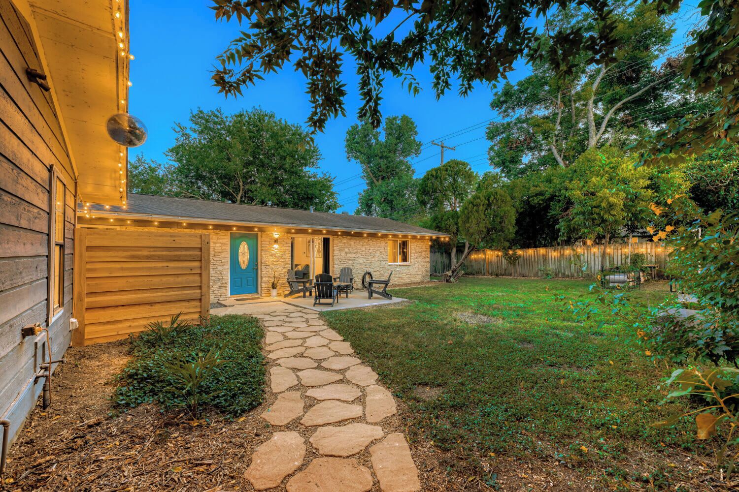 A stone pathway leading to a wooden house with a blue door, outdoor seating, and a fenced backyard.