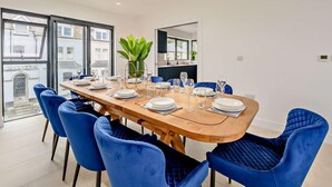 A modern dining area with a wooden table set for six, surrounded by blue chairs, large windows, and a skylight. - 6 Bed in Bideford (oc-91693) (Westward Ho!)