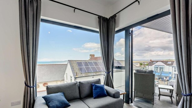 A modern living room with a grey sofa, a small wooden table, and a view of the sea and buildings. - 6 Bed in Bideford (oc-91693) (Westward Ho!)
